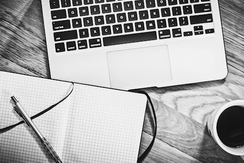 A black-and-white photo of an open notebook, pen, and cup of coffee in front of a laptop.